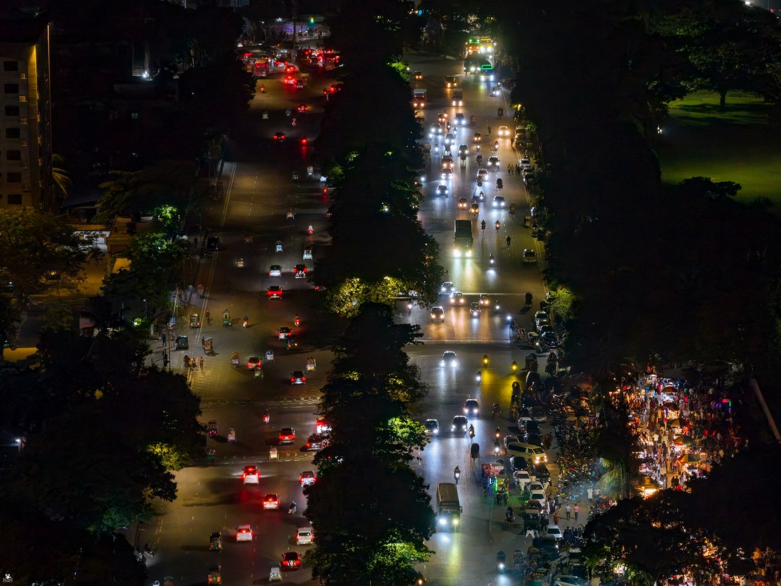 Aerial shot of a lively urban street at night with illuminated traffic and tree-lined roads.