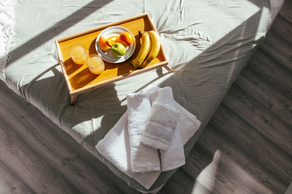 A sunlit bedroom featuring a fresh fruit breakfast tray and neatly folded towels on a soft bed.