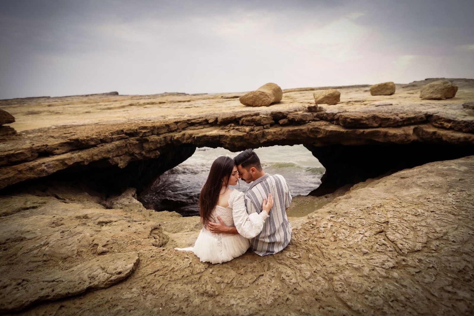 A romantic couple embraces on rocky terrain by the sea under a cloudy sky.