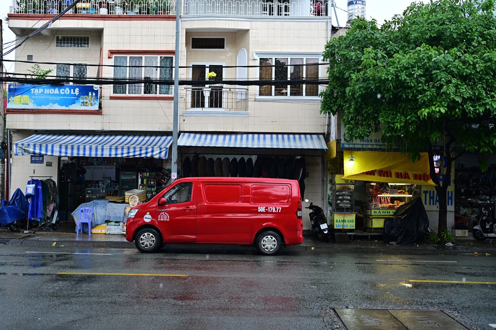 Red van parked outside shops in a rainy Vietnamese street scene.