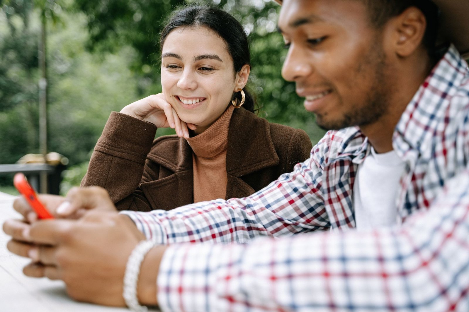 Two friends smiling and enjoying time together outdoors while using a smartphone.