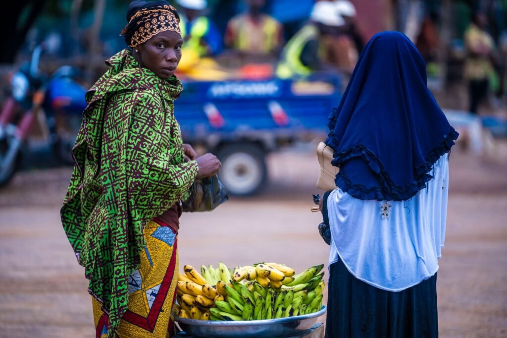 Two women in traditional attire engage in a vibrant African market scene with bananas.