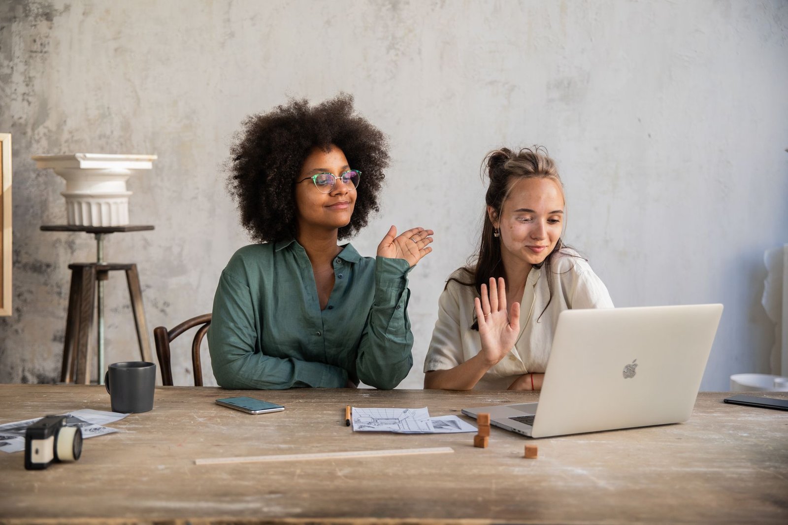 Two diverse women smiling and waving during a video call inside a modern workspace.