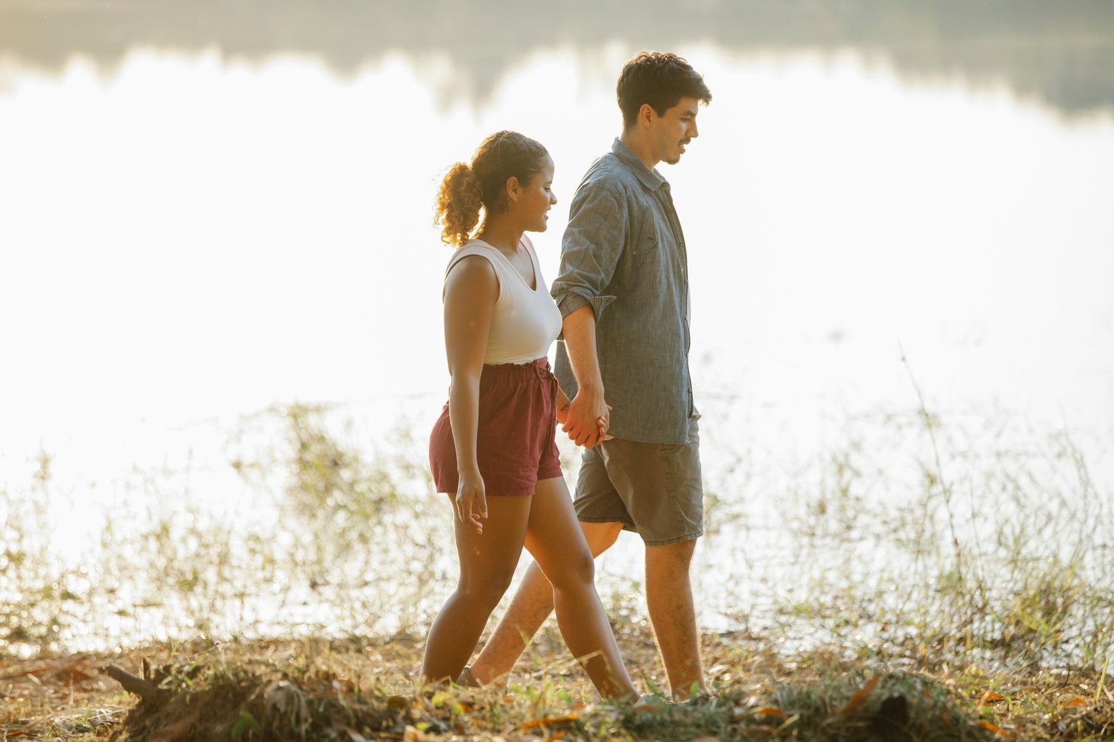 Side view of beloved young multiracial couple walking and holding hands during romantic date near lake at sunset