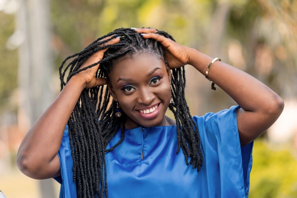 A joyful woman in a blue blouse smiling and holding her braided hair outdoors in Ghana.