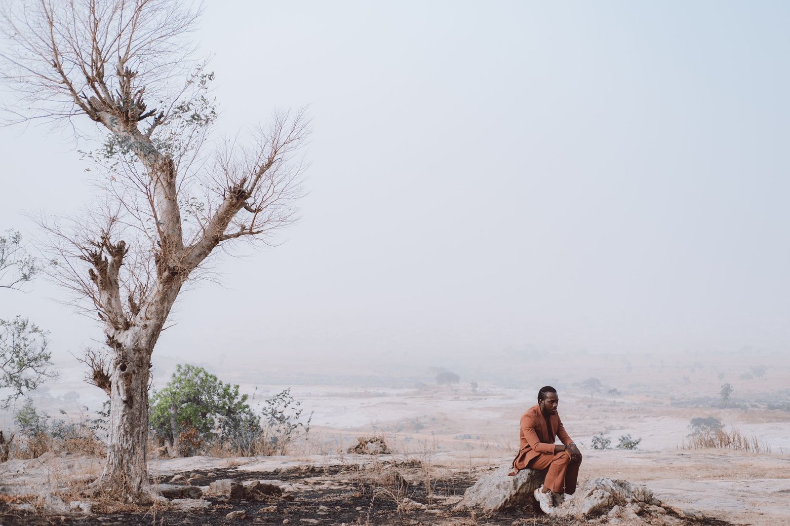 A solitary man in a warm coat sits on a rock under a bare tree in a vast, tranquil winter landscape.