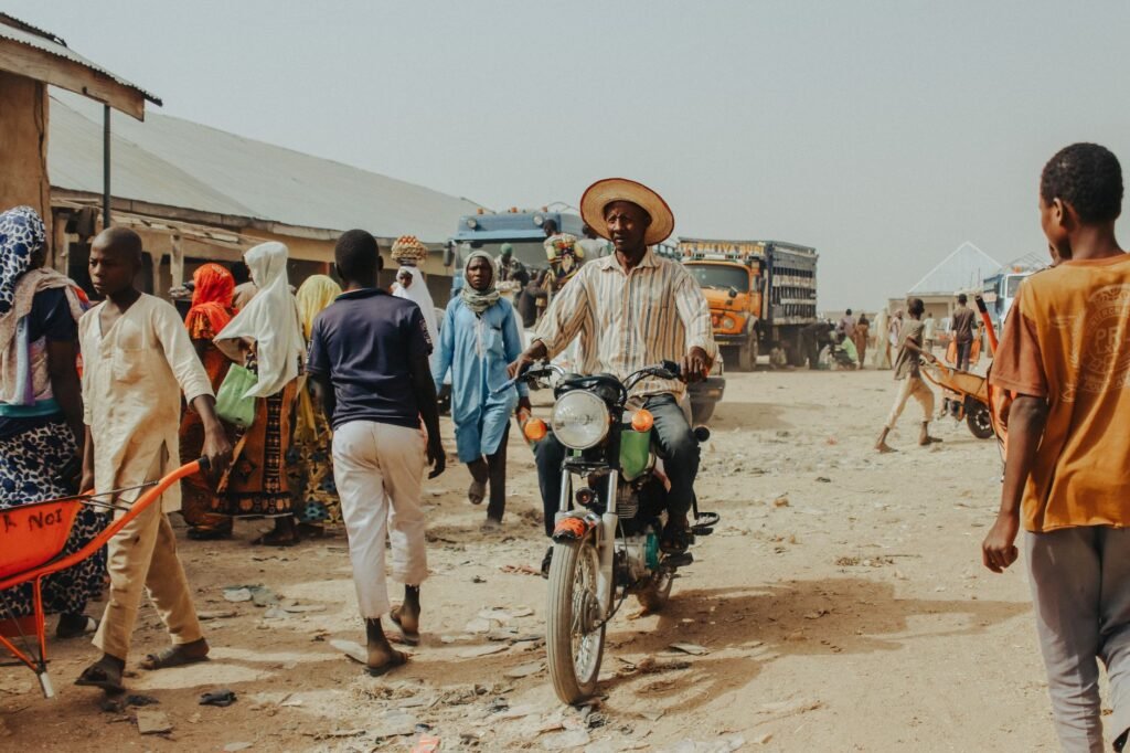 A vibrant village scene with people walking and a man riding a motorbike on a dirt road.