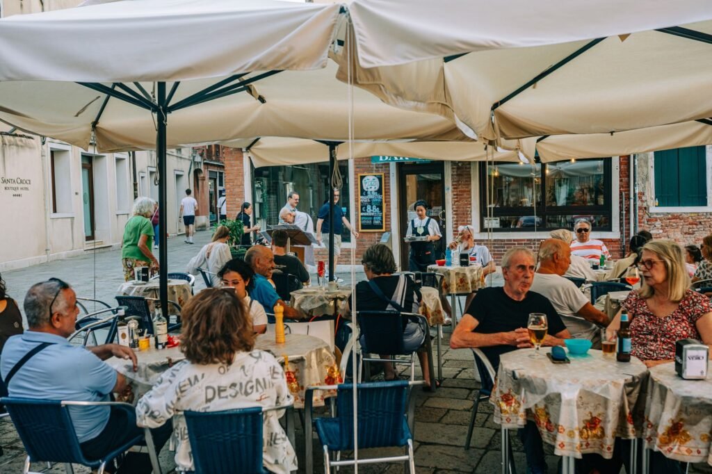 Lively outdoor cafe with people enjoying meals and drinks under umbrellas on a bustling European street.