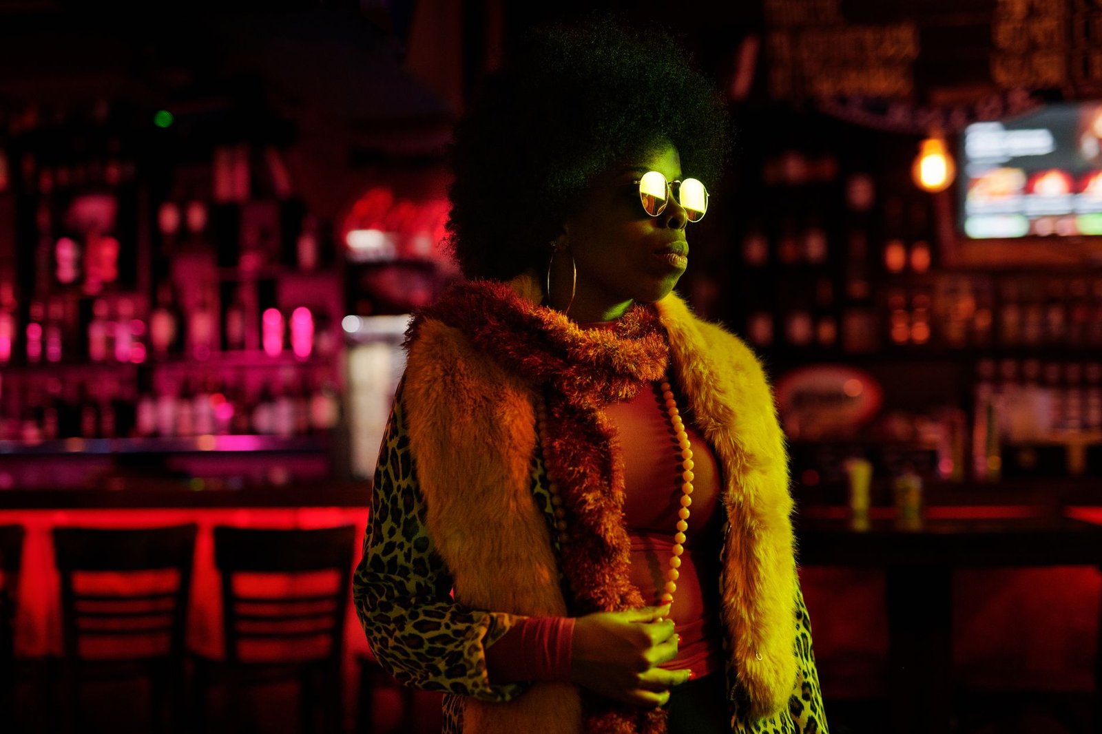 A fashionable woman in a vibrant coat and sunglasses stands in a lively bar at night.