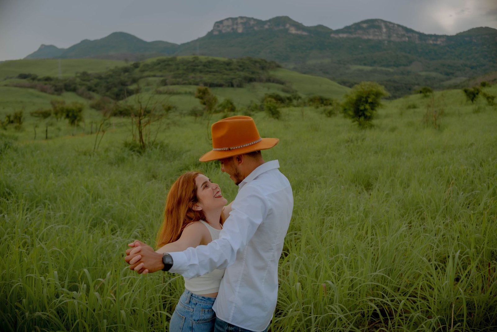 A couple enjoys a romantic moment, embracing in a lush green field with mountain views.