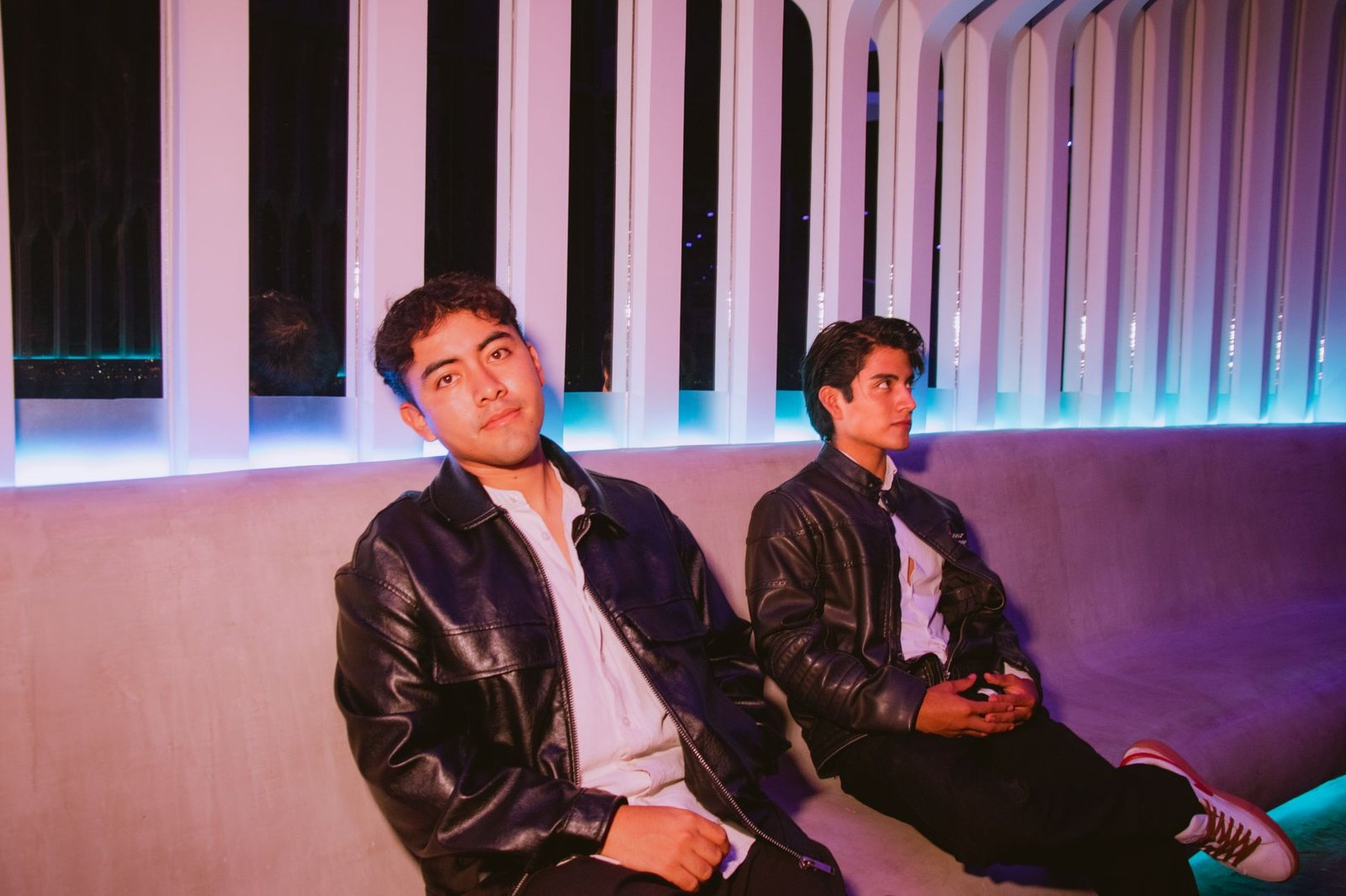 Two young men in leather jackets sit in a modern lounge with neon lighting, creating a stylish and moody atmosphere.