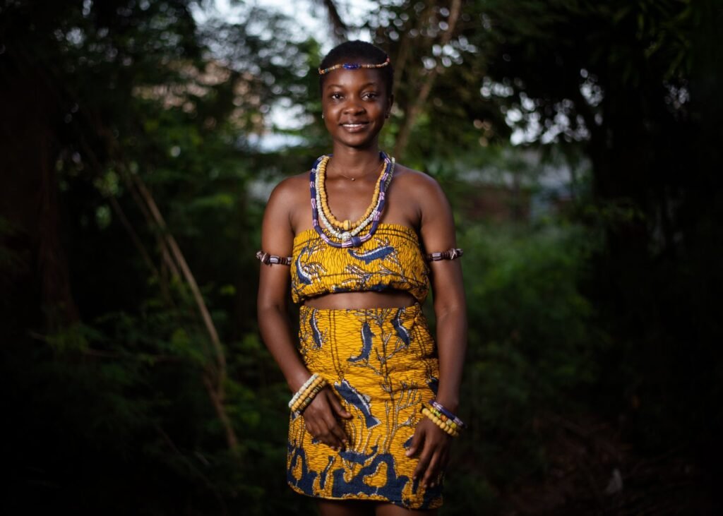 Smiling African woman in traditional yellow attire posing outdoors in a forest setting.