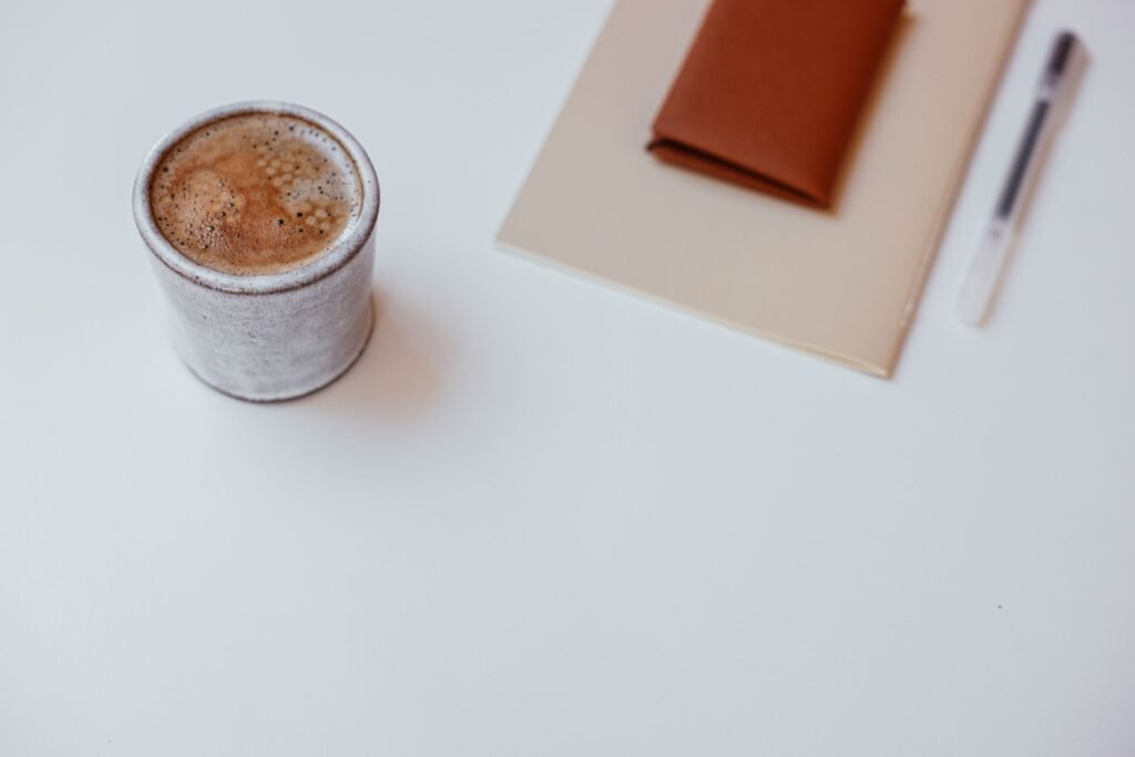Notebook and pen on a table beside a cup of tea with a laptop in the background