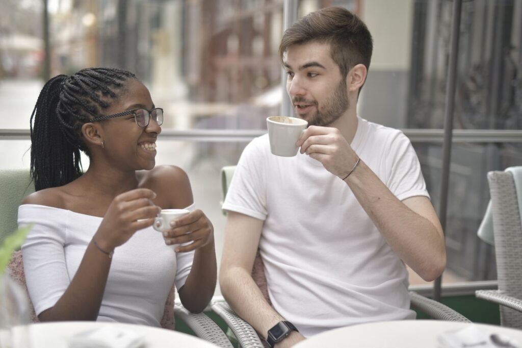Positive young African American woman in eyeglasses sitting near happy boyfriend while drinking coffee together and looking a